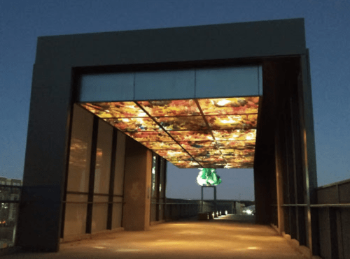 A modern walkway with a colorful ceiling and a green sculpture, illuminated at dusk.