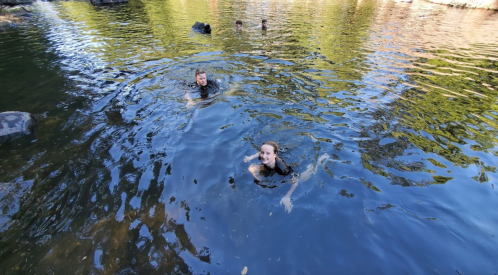 Two people swim in a calm, reflective body of water surrounded by trees, with others visible in the background.