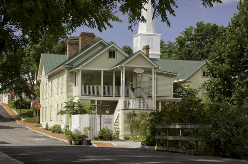 A charming green house with a porch, surrounded by trees, on a quiet street with a church steeple in the background.
