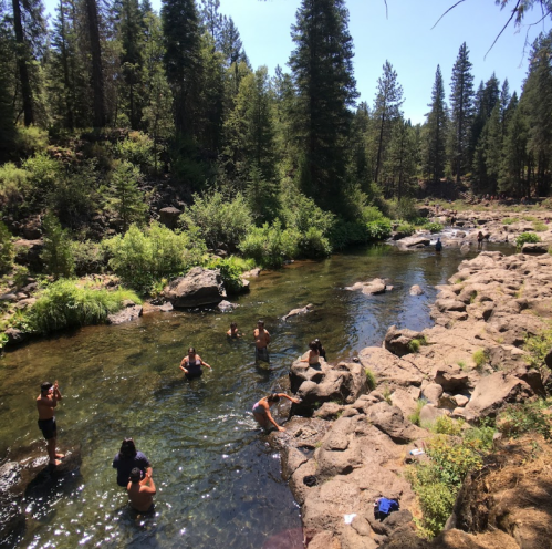 People enjoying a sunny day by a clear stream, surrounded by trees and rocky terrain.