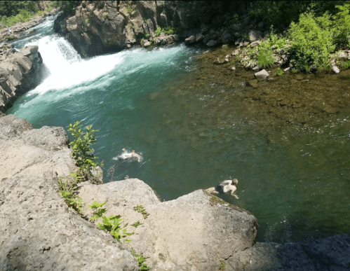 Two people swimming in a clear river surrounded by rocky cliffs and lush greenery, with a waterfall in the background.