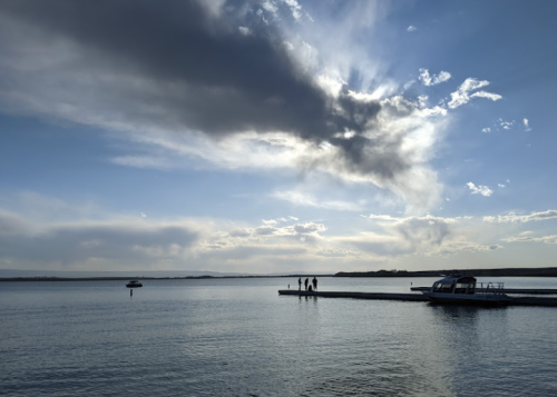 A serene lake scene with a dock, a boat, and silhouettes of people against a cloudy sky.