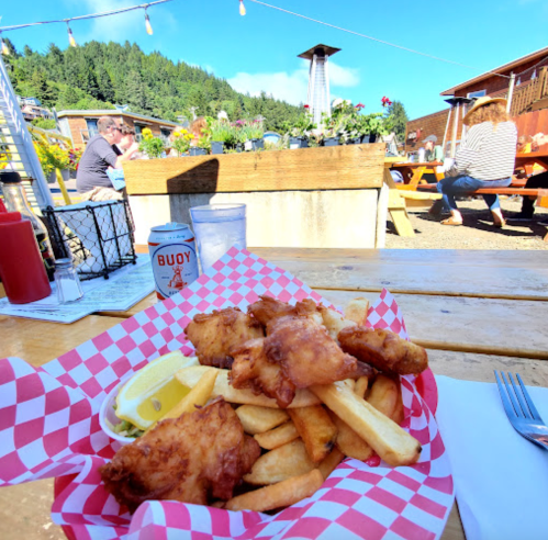 A basket of fish and chips with lemon sits on a wooden table, surrounded by a sunny outdoor dining area.