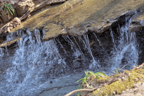 A close-up of a small waterfall cascading over rocky terrain, surrounded by earthy tones and greenery.