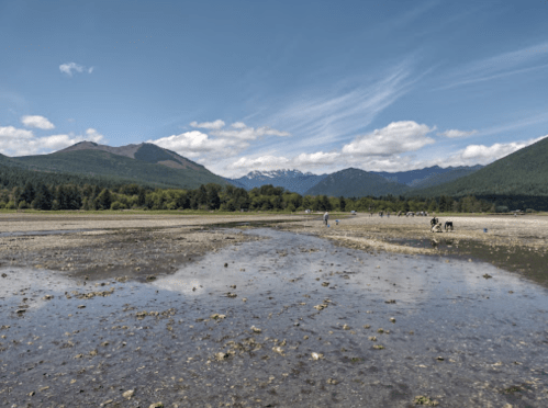 A scenic landscape featuring mountains, a riverbed, and people exploring the area under a clear blue sky.