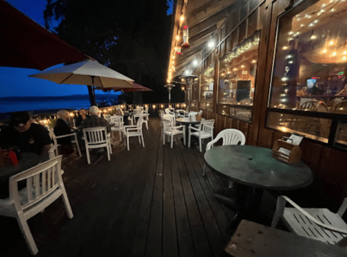 A cozy outdoor dining area with white chairs and tables, illuminated by string lights, near a waterfront at dusk.