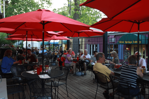 Outdoor dining area with red umbrellas, filled with people enjoying meals at tables surrounded by greenery.
