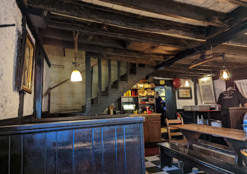 Interior of a rustic restaurant with wooden beams, a staircase, and a dining area featuring wooden tables and chairs.