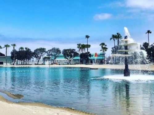 A serene beach scene featuring a fountain in a calm lagoon, surrounded by palm trees and a clear blue sky.