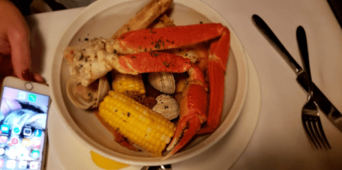 A bowl of seafood featuring crab, clams, corn, and seasonings, with a hand holding a smartphone nearby.