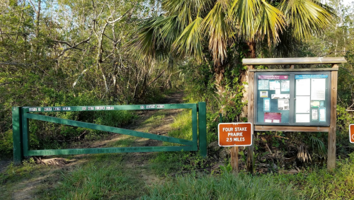 A green gate leads to a trail, with a signboard displaying information and a distance marker for Four Stake Prairie.