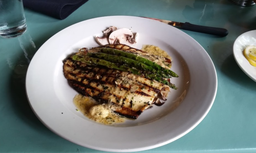 Grilled fish fillet topped with asparagus and mushrooms, served with a buttery sauce on a white plate.