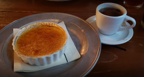 A creamy dessert in a ramekin with a caramelized top, served alongside a small cup of coffee on a metal plate.