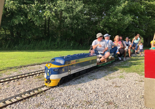 A miniature train with passengers rides along a track in a green park setting. Trees line the background.