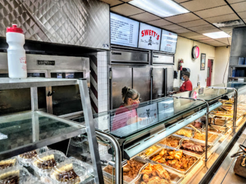 A busy restaurant counter with a glass display of food, staff serving customers, and a menu board above.