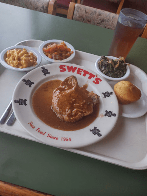 A plate of Southern food featuring pork chop with gravy, macaroni and cheese, greens, cornbread, and a side of peach cobbler.