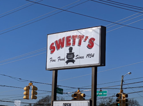 Sign for Swett's restaurant, featuring "Fine Food Since 1954" with a train graphic, under a clear blue sky.