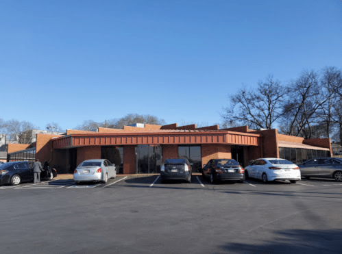 A brick building with a metal roof, surrounded by parked cars and trees under a clear blue sky.
