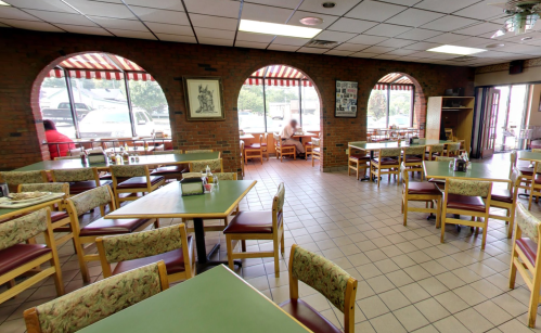 A spacious restaurant interior with empty tables, brick walls, and large windows, creating a bright atmosphere.