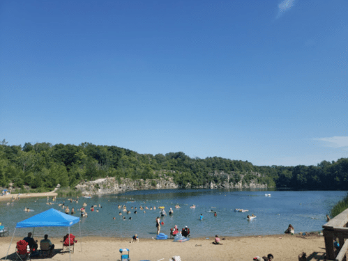 A sunny day at a lake with people swimming and relaxing on the beach, surrounded by green trees and cliffs.