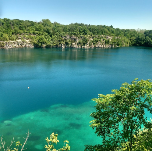 A serene blue lake surrounded by lush green trees and rocky cliffs under a clear blue sky.