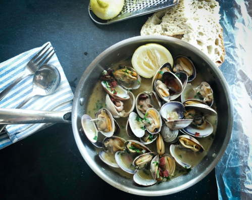 A pan of cooked clams with herbs and lemon, accompanied by bread and utensils on a dark countertop.