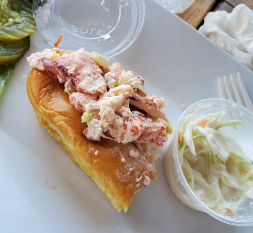 A lobster roll on a bun topped with lobster salad, served with a side of coleslaw and pickles.