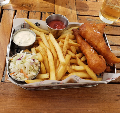 A basket of fish and chips with two fried fish fillets, fries, coleslaw, and dipping sauces on a wooden table.