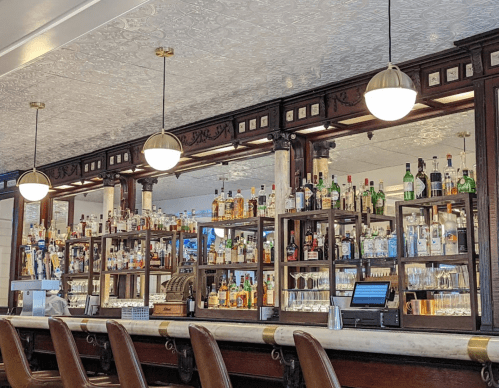 A stylish bar with a wooden counter, shelves filled with various liquor bottles, and elegant pendant lights.