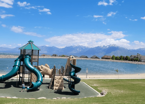 A playground with slides near a lake and mountains under a blue sky with scattered clouds.
