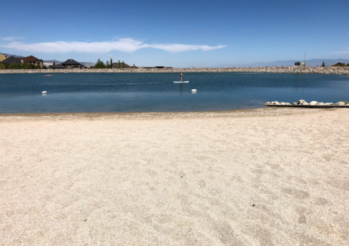 A sandy beach by a calm lake, with a paddleboarder in the distance and mountains under a clear blue sky.