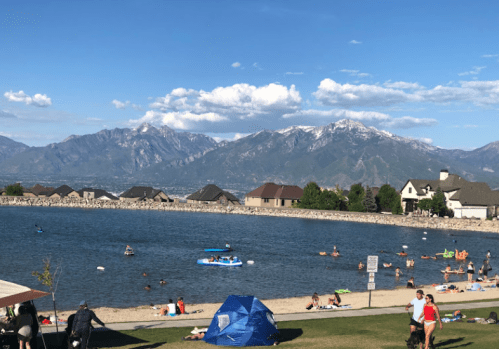 A scenic view of a lake with people swimming and kayaking, surrounded by mountains and houses under a clear blue sky.