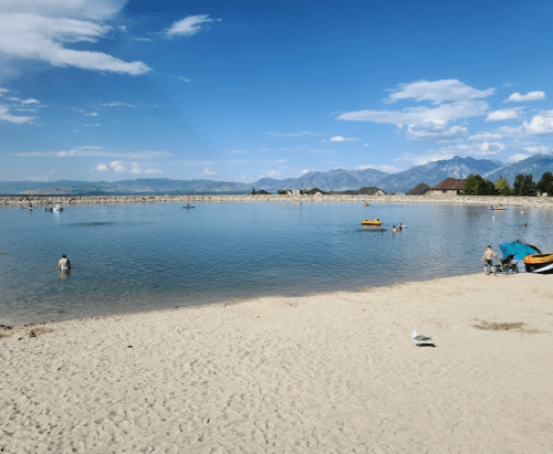 A sandy beach by a calm lake with people swimming and mountains in the background under a clear blue sky.
