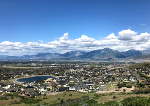 A panoramic view of a valley with houses, a pond, and mountains under a blue sky with scattered clouds.