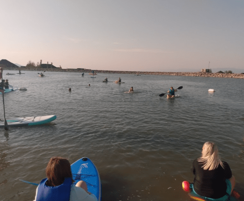 People kayaking and paddleboarding on a calm water body during sunset, with a rocky shore in the background.