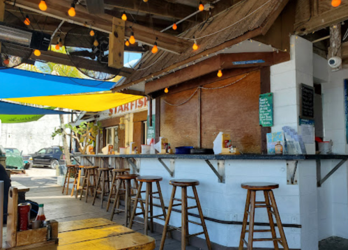 A rustic outdoor bar with wooden stools, colorful canopies, and string lights, set in a casual dining area.