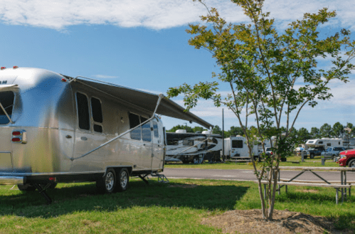 A shiny silver Airstream trailer parked in a grassy area with trees and other RVs in the background under a blue sky.