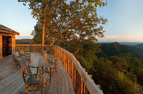 A wooden deck with chairs overlooks a lush green mountain landscape at sunset.