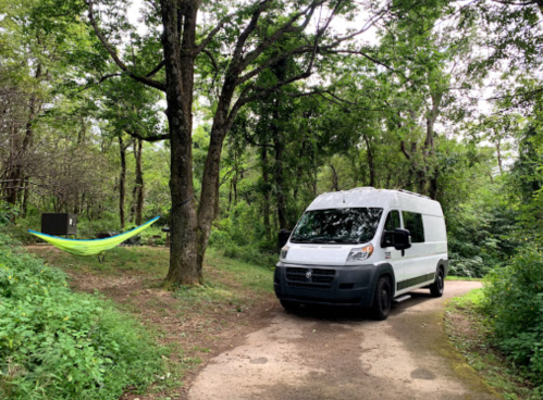 A white van parked on a dirt path in a wooded area, with a hammock hanging between trees nearby.