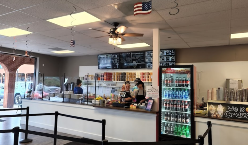A bright ice cream shop interior with a service counter, menu board, and a variety of drinks in a cooler.