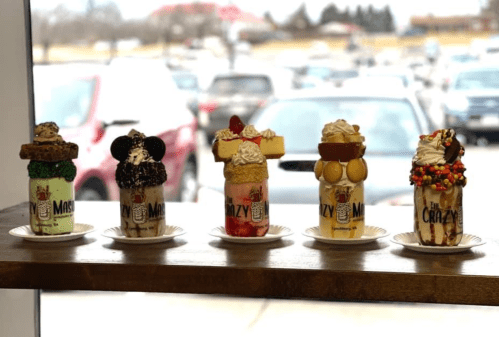 Five colorful milkshakes topped with various sweets, displayed on a wooden ledge with cars in the background.
