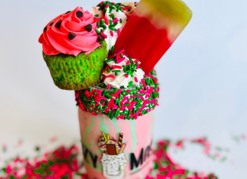 A colorful milkshake topped with a cupcake, sprinkles, and a popsicle, all in a decorated glass.