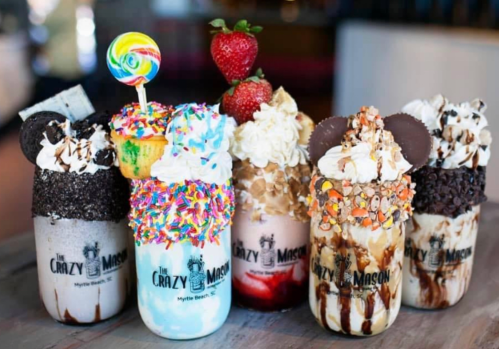 Five colorful milkshakes topped with candy, whipped cream, and fruit, served in mason jars with fun decorations.