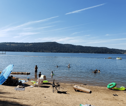 A sunny beach scene with people swimming, lounging, and enjoying the water near a lake surrounded by trees.
