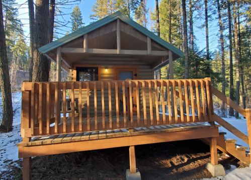 A cozy cabin with a wooden deck, surrounded by tall trees and snow, under a clear blue sky.