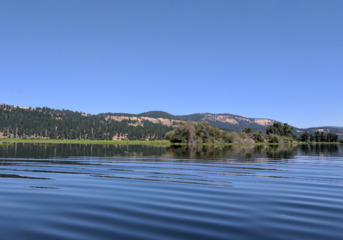 A serene lake surrounded by lush greenery and distant hills under a clear blue sky. Gentle ripples on the water's surface.