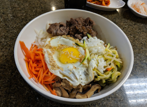 A bowl of mixed ingredients featuring rice, vegetables, beef, and a fried egg on top.