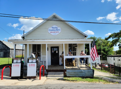 A small country store with an American flag, featuring two people sitting outside on a sunny day.