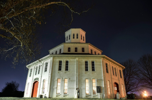 Historic round building illuminated at night, surrounded by bare trees against a starry sky.
