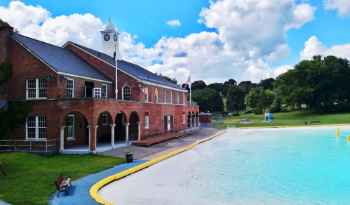 A brick building with a clock tower beside a clear blue pool and green park under a partly cloudy sky.